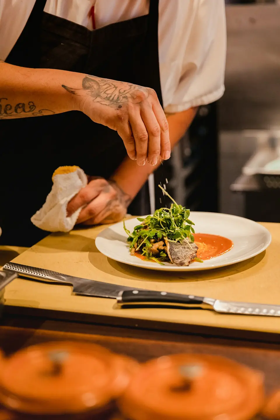 Les mains du chef déposent des herbes fraîches sur une assiette, devant des cocottes en fonte orange
