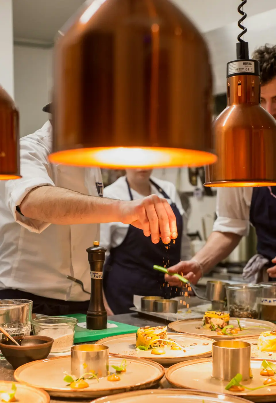 Cuisiniers en tablier assaisonnant des assiettes sous des lampes chauffantes en cuivre