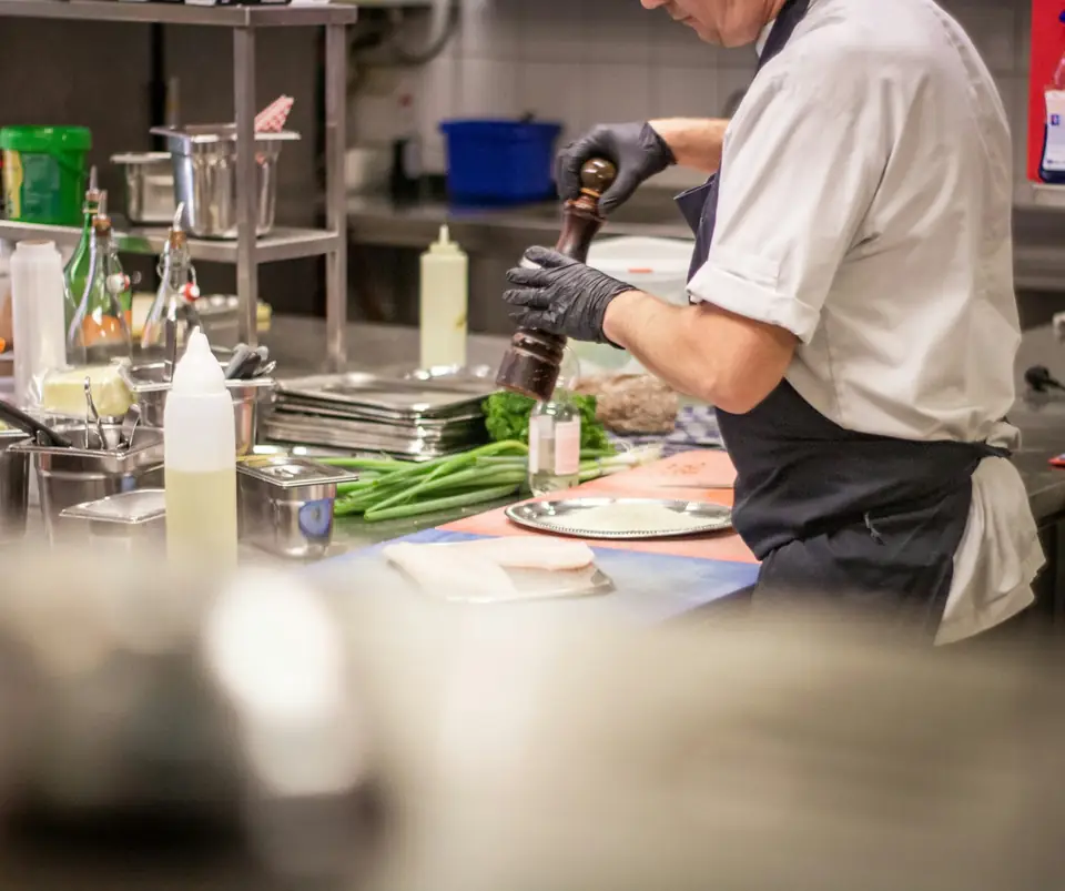 Cuisinier en tablier noir et gants donnant un tour de moulin à poivre au-dessus du plan de travail