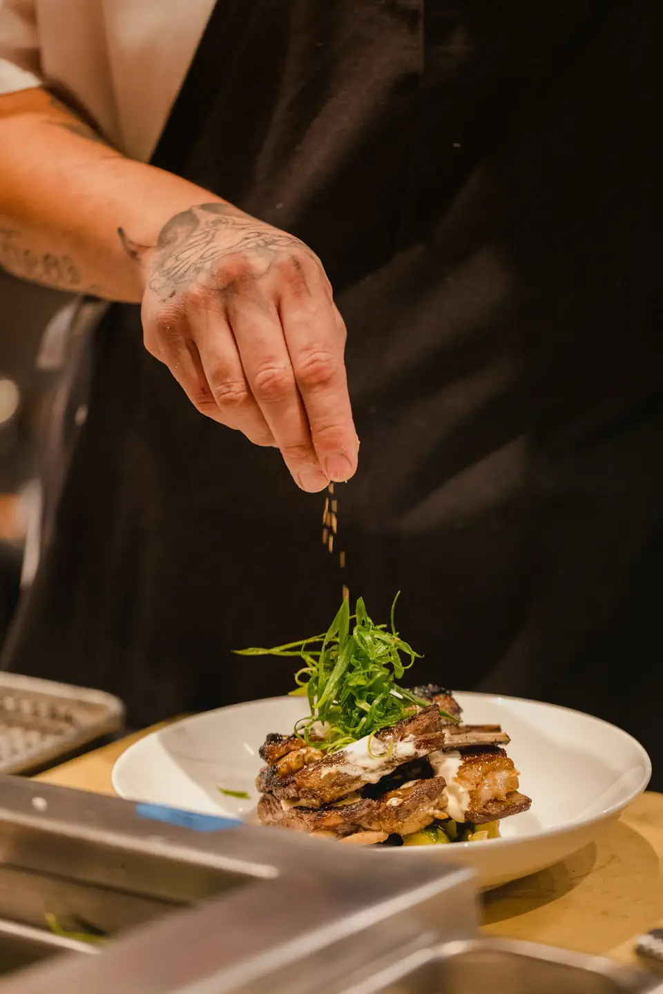 Main déposant une pincée d’herbes sur une assiette, sur fond sombre