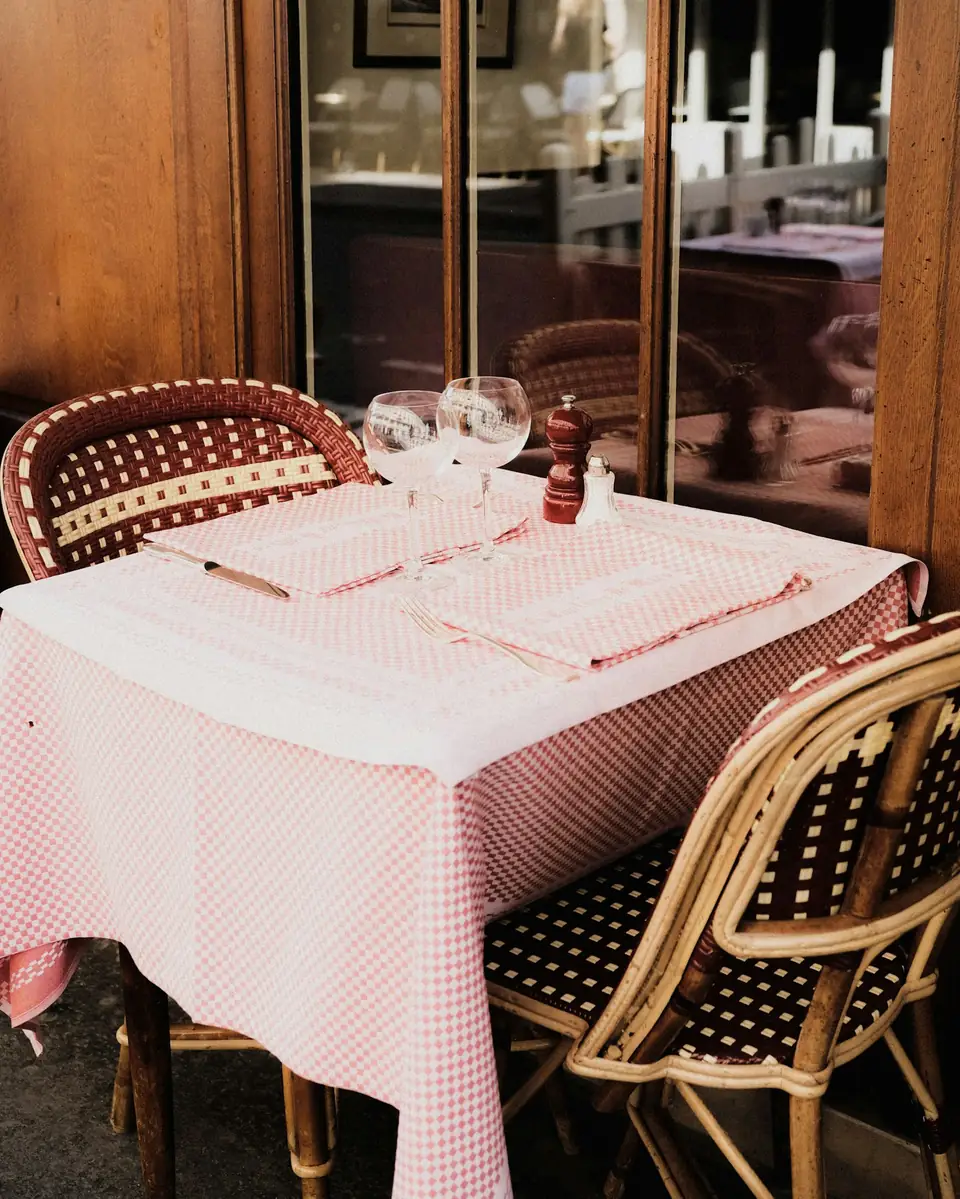 Table dressée avec une nappe vichy rose et des chaises en rotin tressé, près d’une porte vitrée